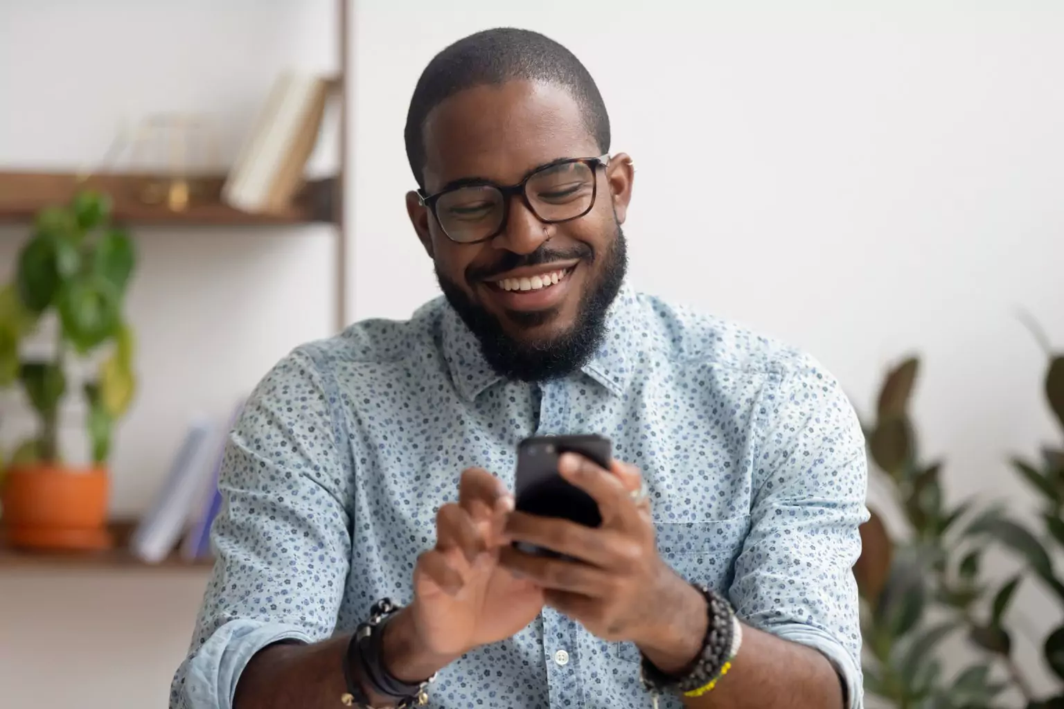 Man in glasses smiling as he reads his phone at a desk by a window